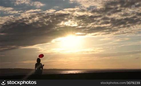 Back view of sad lonely charming female holding red heart shaped balloon, sitting on seashore and watching sunset. Pensive broken hearted girl with balloon sitting alone on seaside in rays of orange setting sun on windy summer day. Slow motion.