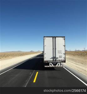 Back view of semi truck traveling down highway towards horizon under clear blue sky.