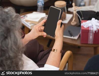 Back view of senior elder asian grandmother holding smart phone tablet sit on sofa or couch in living room at home.