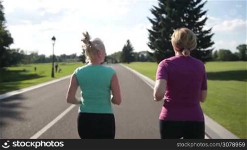 Back view of senior female joggers running on the road in park. Rear view of adult blonde women in sport clothes jogging on city park path on sunny day. Slow motion. Steadicam stabilized shot.
