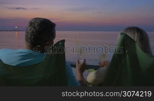 Back view of senior man and woman relaxing in chaise-longues on thee bach in late evening. They toasting with cocktails and enjoying seascape