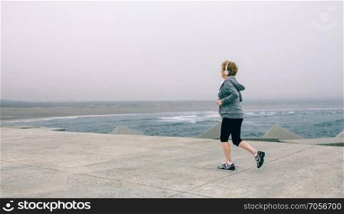Back view of senior sportswoman with headphones running by sea pier. Senior sportswoman with headphones running
