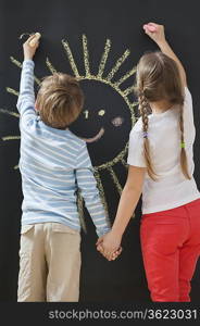 Back view of siblings drawing sun on blackboard while holding hands