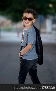 Back view of stylish rocker in sunglasses holding jacket on shoulder and smiling over shoulder.. Charming trendy boy posing at camera