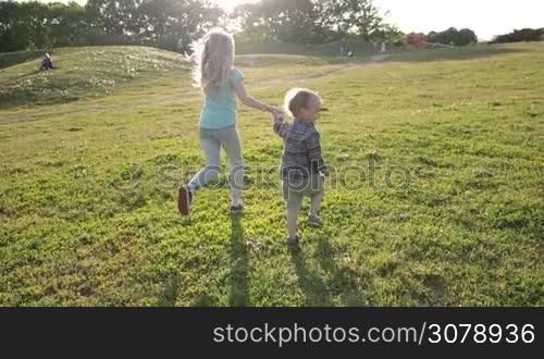 Back view of two playful children holding hands running through green grassy field in park. Young sister and toddler brother having fun and running on green meadow in glow of sunset. Slow motion. Steadicam stabilized shot.