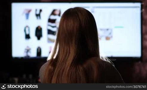 Back view of woman&acute;s sihlouette sitting in front of wide computer display and browisng websites. Woman shopping online at home using pc with wide display, blurred online shops on computer&acute;s screen.