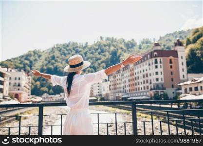 Back view of woman in hat on the embankment of a mountain river in a European city. Happy summer travel concept. Rosa Khutor, Mzymta River, Sochi, Russia. Happy girl at hat on the embankment of a mountain river in a European city.