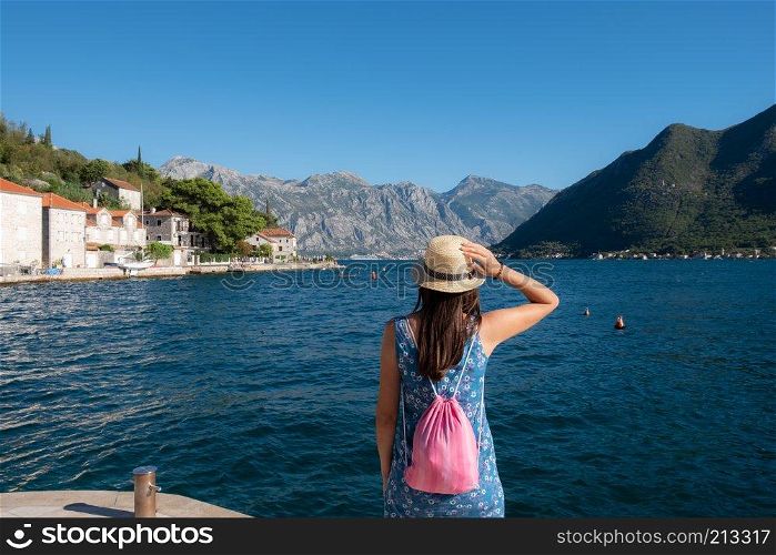 Back view of woman in hat watching the sea with the mountains and Mediterranean town at the background, Perast, Montenegro