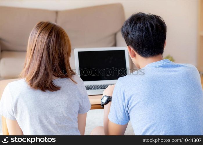 Back view of young asian couple working laptop with blank screen display, family planning and searching content together, man and woman looking computer, business and communication concept.