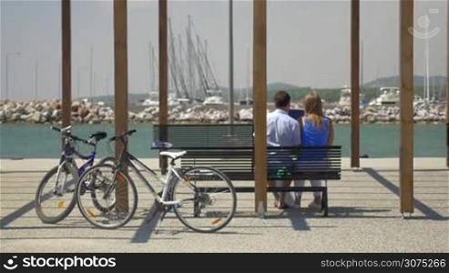 Back view of young couple with tablet computer sitting on the bench on water front, parked bicycles nearby. Yachts in background