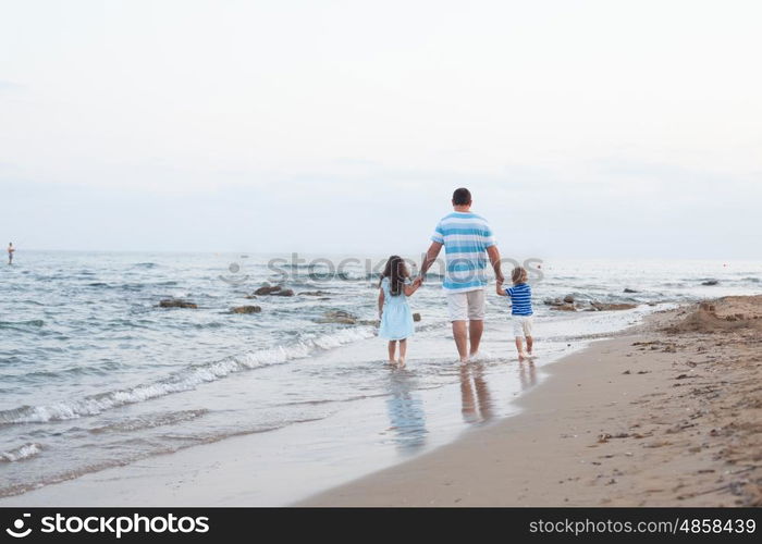 Back view of young father and his two pretty kids walking along sand beach at evening time. Walking on the empty beach