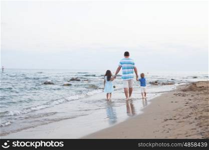 Back view of young father and his two pretty kids walking along sand beach at evening time. Walking on the empty beach