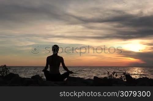 Back view of young female practicing yoga, sitting in lotus position on the beach during sunset. Silhouette of yoga woman meditating on seaside over red sky with clouds at sundown. Slow motion. Steadicam stabilized shot.
