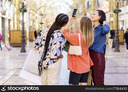 Back view of young multiracial female friends wearing trendy outfits standing with shopping bags and taking picture on smartphone on pedestrian street. Stylish diverse women standing on street and taking photo