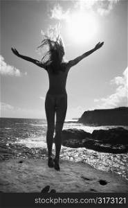 Back view of young nude Asian woman jumping into the air facing the ocean.