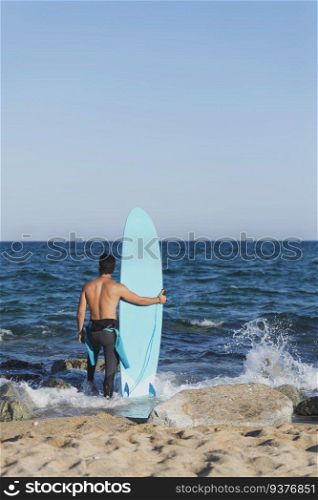 Back view of young surfer standing barefooted on sandy shore, facing vast ocean and holding his surfboard, ready to hit waves on windy summer day