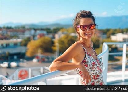 Back view of young woman standing on the deck of the ship or ferry boat in sunny day on vacation traveling wearing summer dress looking to the camera smiling wearing sunglasses