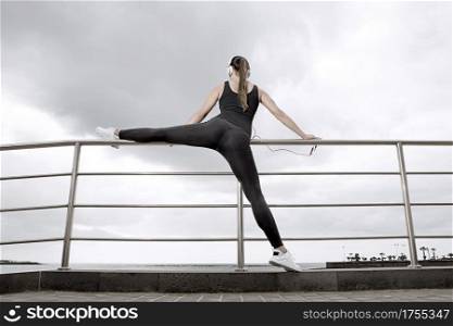 Back view of young woman stretching the leg and listening to music. Horizontal outdoors shot. . Female warming up before training