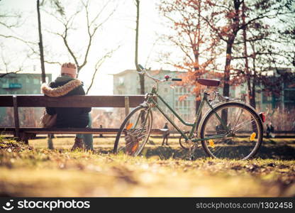 Back view of young woman with bicycle who is enjoying the sun on a park bench, spring time