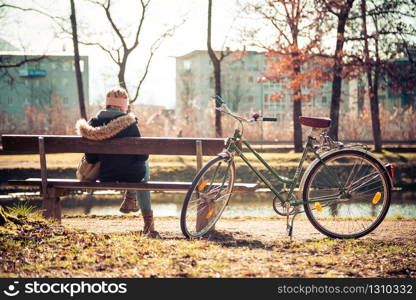 Back view of young woman with bicycle who is enjoying the sun on a park bench, spring time