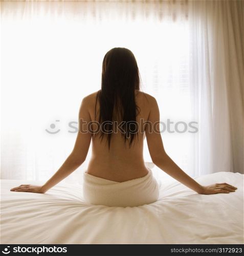 Back view portrait of pretty Caucasian young woman partially nude sitting on bed by sunlit window.