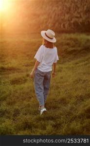 Back view portrait of young woman walking across meadow on summer sunny day, wearing straw hat lit by sunset light, copy space, back view.. Back view portrait of young woman walking across meadow on summer sunny day, wearing straw hat lit by sunset light, copy space, back view