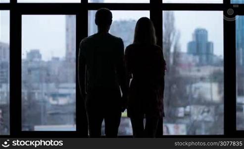 Back view silhouettes of two lovers looking thoughtfully out of the window. Slow motion. Rear view of affectionate couple standing by wide window and cuddling each other, loving woman putting her head onto man&acute;s shoulder.