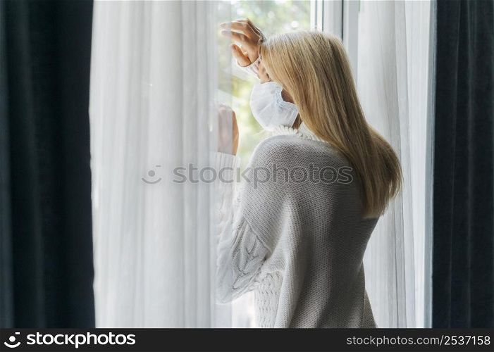 back view woman with medical mask home during pandemic looking through window