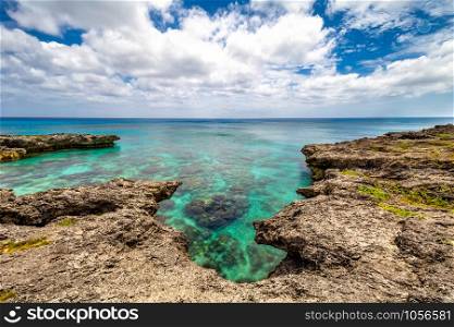 Background view of shallow turquoise waters with coral reefs underneath the surface and fringing reefs encircling it above the surface. Clear horizon line, blue sky and clouds in the background.