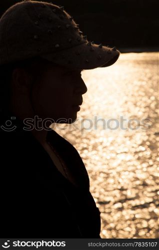 Backlit silhouette of a woman wearing a hat. Sop freckled sunlight reflecting off the surface of the sea in the evening.