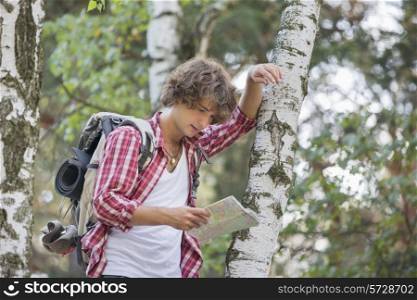 Backpacker reading map while leaning on tree trunk in forest
