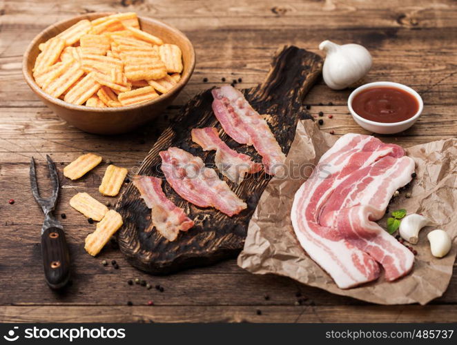 Bacon flavored snacks chips with grilled bacon rashers on vintage chopping board with smoked raw bacon on butchers paper with garlic and sauce on wood background.