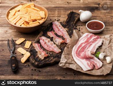 Bacon flavored snacks chips with grilled bacon rashers on vintage chopping board with smoked raw bacon on butchers paper with garlic and sauce on wood background.