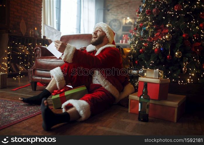 Bad drunk Santa claus reads letters under christmas tree, nasty party, humor. Unhealthy lifestyle, bearded man in holiday costume, new year. Bad drunk Santa claus reads letters, nasty man