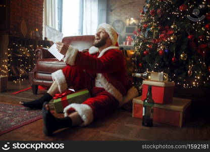 Bad drunk Santa claus reads letters under christmas tree, nasty party, humor. Unhealthy lifestyle, bearded man in holiday costume, new year. Bad drunk Santa claus reads letters, nasty man