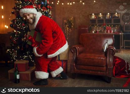 Bad drunk Santa claus steals gifts under christmas tree, nasty party, humor. Unhealthy lifestyle, bearded man in holiday costume, new year and alcoholism. Bad drunk Santa steals gifts under christmas tree