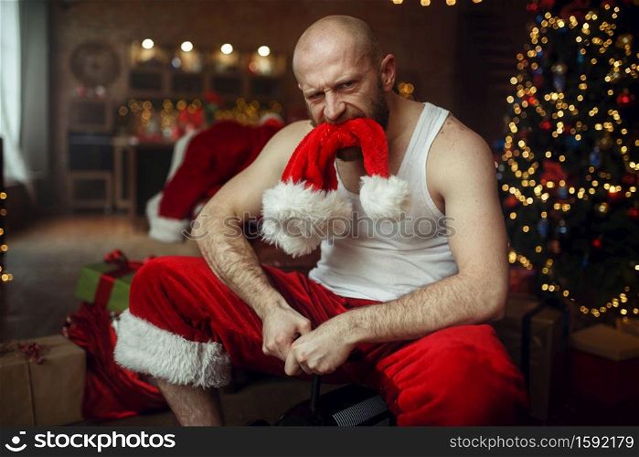 Bad drunk Santa claus with red hat in mouth riding on little toy car, nasty party, humor. Unhealthy lifestyle, bearded man in holiday costume, new year and alcoholism. Bad drunk Santa claus with red hat in mouth
