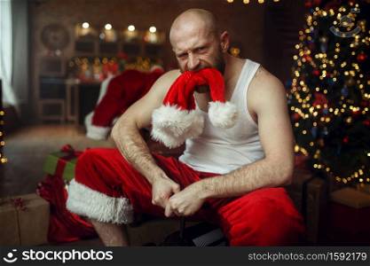 Bad drunk Santa claus with red hat in mouth riding on little toy car, nasty party, humor. Unhealthy lifestyle, bearded man in holiday costume, new year and alcoholism. Bad drunk Santa claus with red hat in mouth