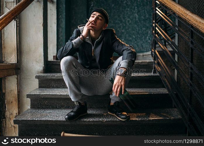 Bad guy with bottle of beer sits on the stairs and smoking. Street robber waiting for victim. Crime concept