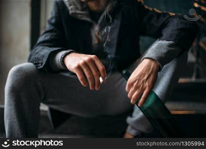 Bad guy with bottle of beer sits on the stairs and smoking. Street robber waiting for victim. Crime concept. Guy with bottle sits on the stairs and smoking