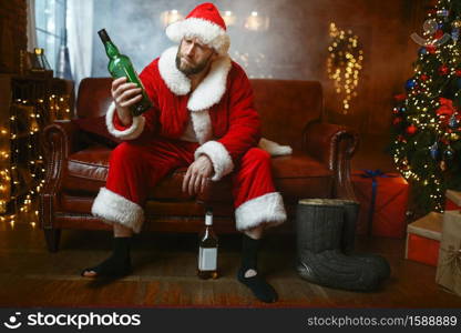 Bad Santa claus celebrate christmas with bottle of alcohol on couch. Unhealthy lifestyle, bearded man in holiday costume, new year and alcoholism. Bad Santa claus celebrate christmas with bottle