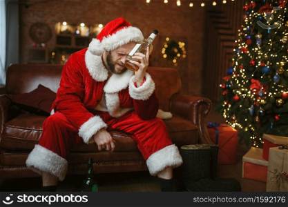 Bad Santa claus with bottle of alcohol sitting on couch, hangover. Unhealthy lifestyle, bearded man in holiday costume, new year and alcoholism. Bad Santa claus with alcohol on couch, hangover