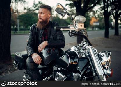 Baerded biker poses on chopper leaning on a helmet. Vintage bike, rider and his motorcycle, freedom lifestyle, biking
