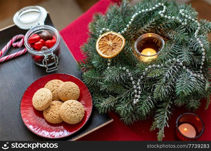bake, food and winter holidays concept - oatmeal cookies on red ceramic plate, chocolate candies and christmas wreath with burning candle at home. cookies, candies and christmas wreath with candle