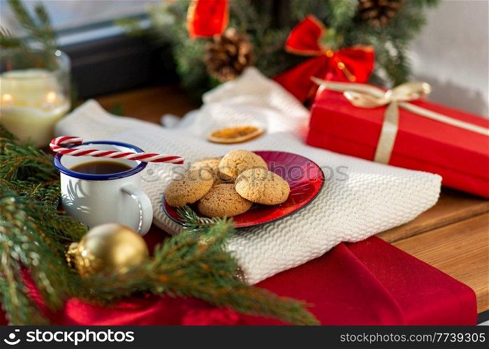 bake, food and winter holidays concept - oatmeal cookies on red ceramic plate, cup of coffee, gift and christmas decorations on window sill at home. cookies, coffee and christmas gift on sill at home