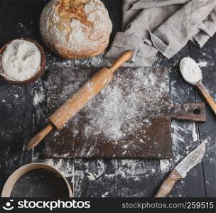 baked bread, white wheat flour, wooden rolling pin and old cutting board on a black table, top view