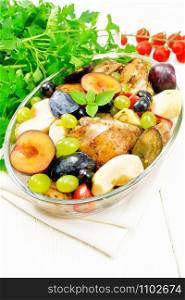 Baked chicken with tomatoes, apples, plums and grapes in a glass roaster on a light napkin, garlic, parsley and basil on white wooden board background