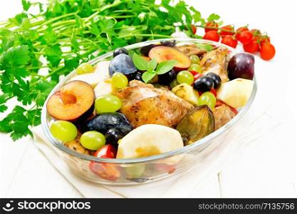 Baked chicken with tomatoes, apples, plums and grapes in a glass roaster on a kitchen towel, garlic, parsley and basil on background of light wooden board