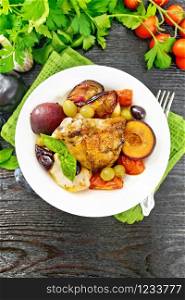 Baked chicken with tomatoes, apples, plums and grapes in a plate on a napkin, garlic, parsley and basil on dark wooden board background from above