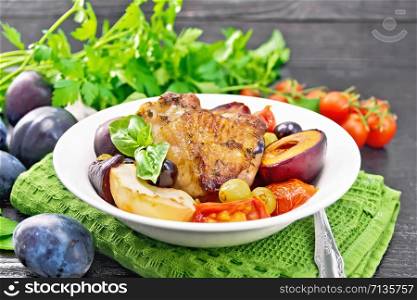 Baked chicken with tomatoes, apples, plums and grapes in a plate on green napkin, garlic, parsley and basil on wooden board background